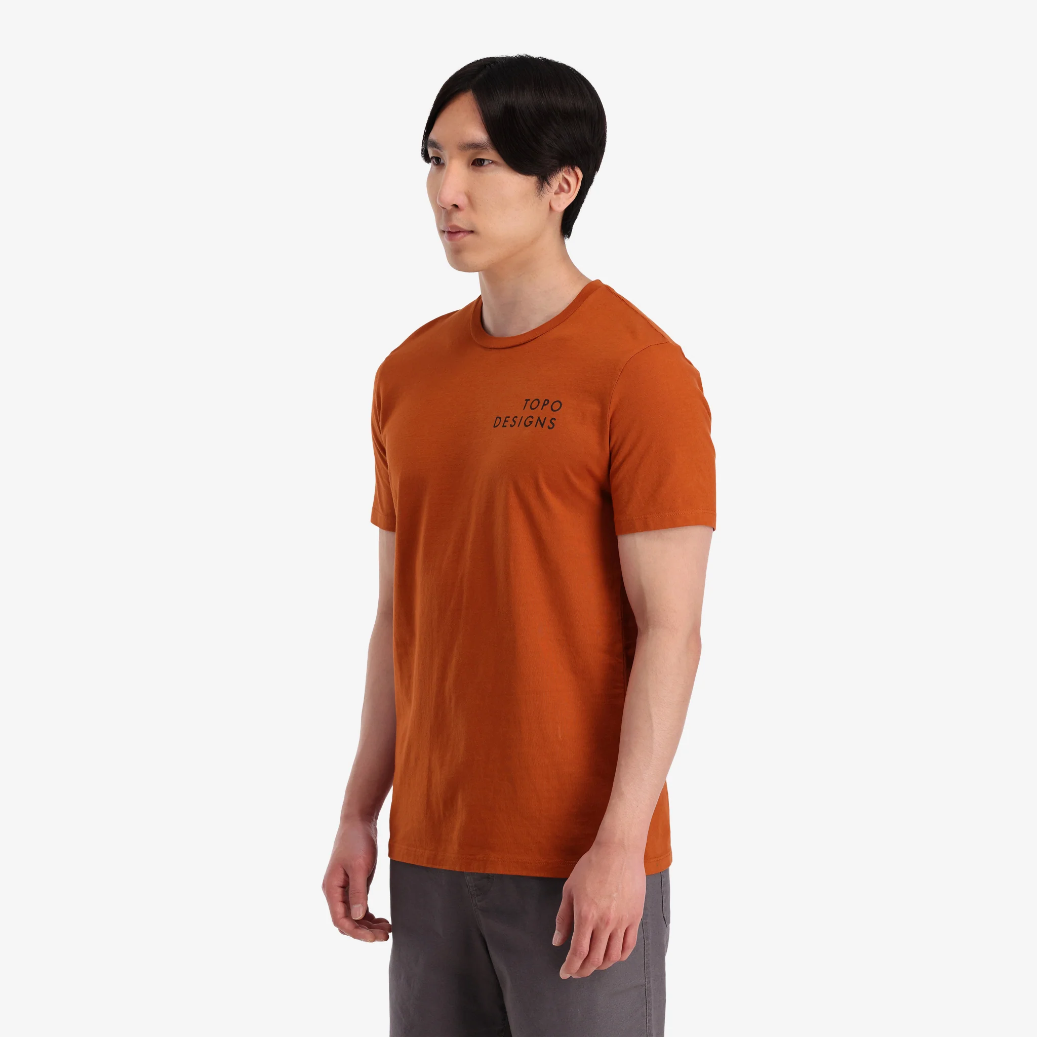 General shot on-model left side view of Topo Designs Men's Camp Logo Tee 100% organic cotton graphic short sleeve t-shirt in "clay" orange.